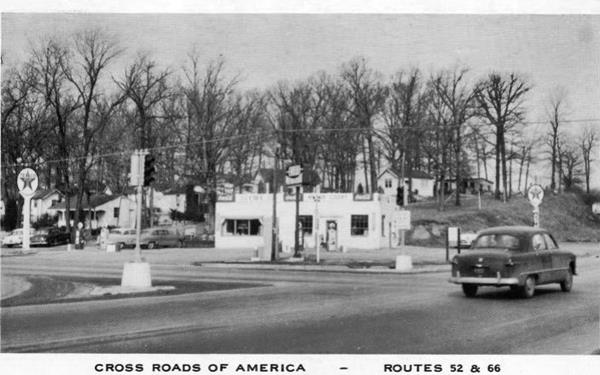 black and white 1950s Texaco on a crossroads, woods and houses beyond. Car on highway 66