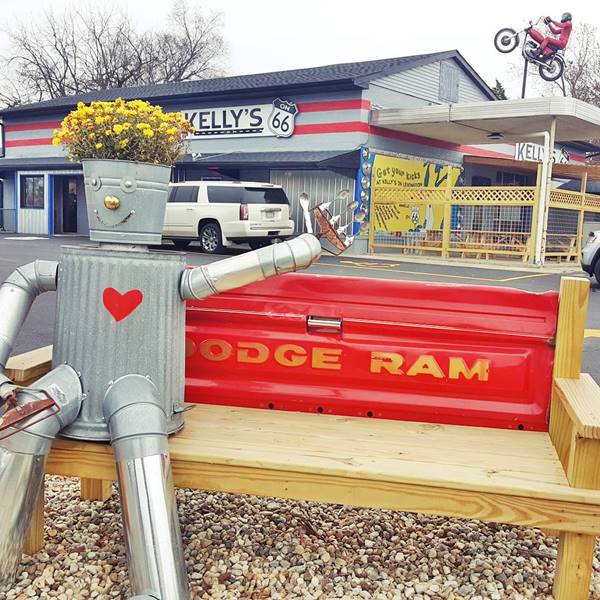 color, bench with a red Dodge Ram tailgate, robot sitting on it, building behind with US66 shield, and a motorcycle doing wheelie atop a post