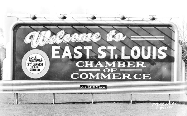 black and white photo from 1940s9 of a park with trees and a 
billboard with the words "Welcome to East St Louis. Nations 2nd largest rail center. Chamber of Commerce" in dark letters on white background