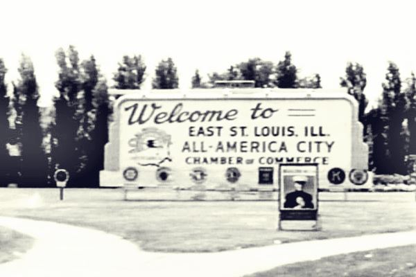 black and white photo from 1959 of a park with trees and a 
billboard with the words "Welcome to East St Louis. ILL. All-America City, Chamber of Commerce" in dark letters on white background