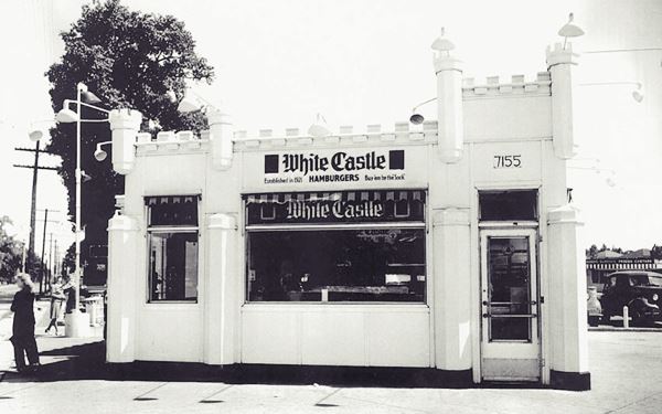 black and white c.1940 single story castle-shaped small building on a corner, cars and woman