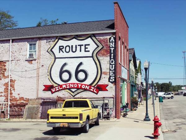 color, mural with US66 shield painted on a red brick wall, yellow pick up truck parked beside it, street to the right