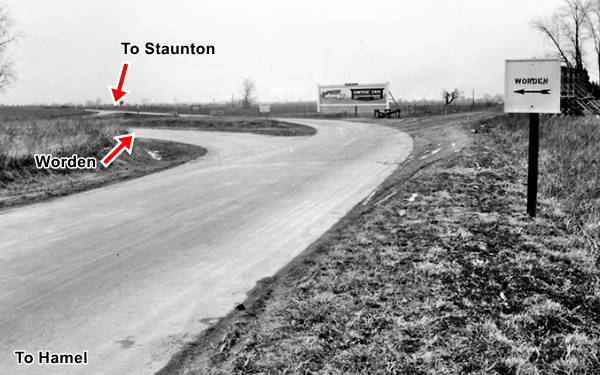 concrete road curves at a junction, in the center, a road leaves to the left. Sign pointing left says "Worden" trees to the right and a billboard. Black and white 1930s photo