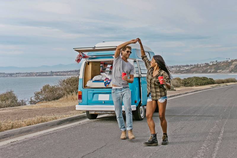 young couple dance by their van with paper cups in their hands on the road next to a lake