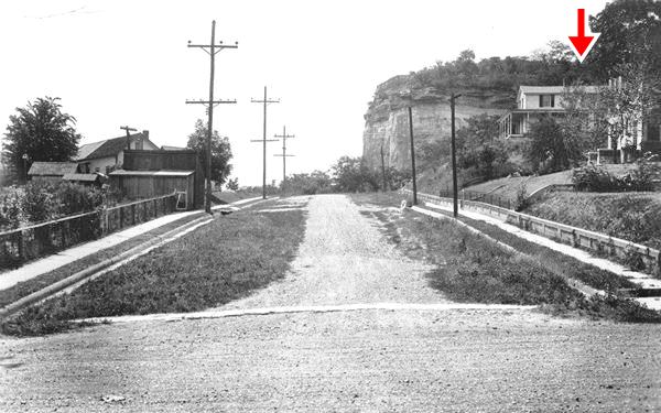 gravel street at the foot of a bluff, home to the right gable roof in a black and white photo from 1930