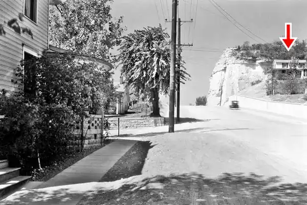 gable roof brick home (right), white bluffs with vegetation behind in it. US 66 rusns by foot of cliff and home, black-white c.1935