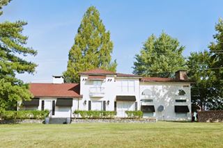 two story hipped and gable red-tile roofed home with trees around it and lawn in front seen from Route 66