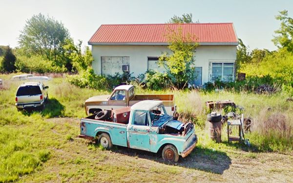 junkyard, rusting cars and trucks in tall grass by Red-roofed Garage