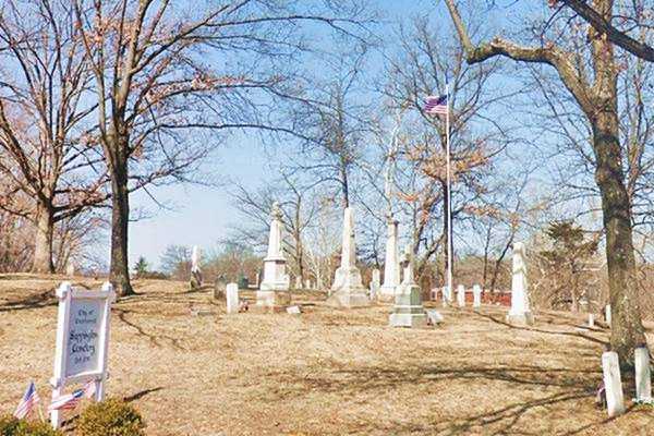 tombstones, grave memorials and an American flag, with a lawn and trees in March