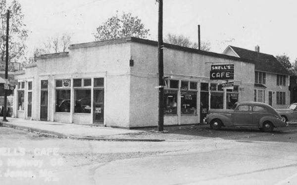 black and white photo c. 1930s of one story stucco walled corner building and a 1950s car, a bar on Route 66