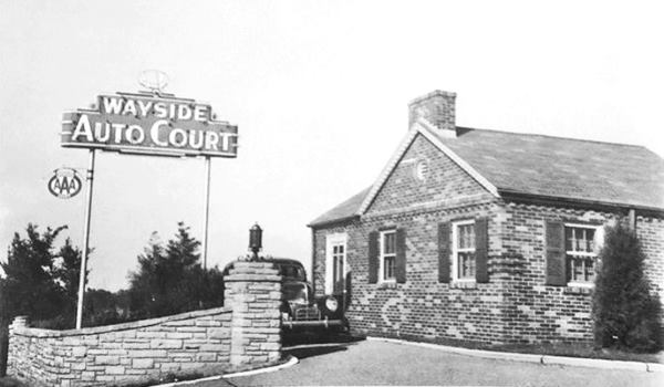 stone wall, car and brick building with gable roof and motel neon sign in a black and white 1930s photo