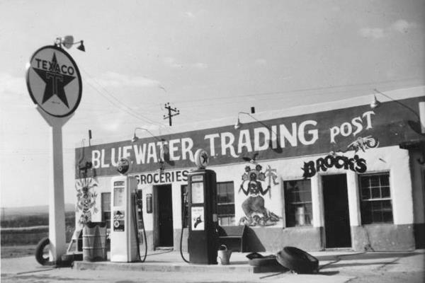 black and white 1948 photo: left Texaco neon sign, block-shaped trading post with murals 2 gas pumps and some tires