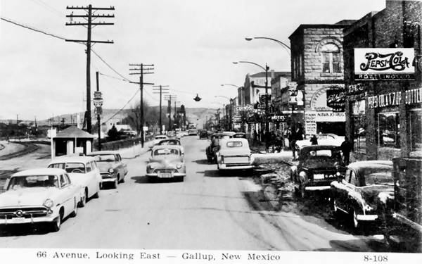 black and white mid 1950s view east along Route 66, narrow street. Buildings with neon signs on right, trees along left and railroad tracks. Cars driving both ways and parked