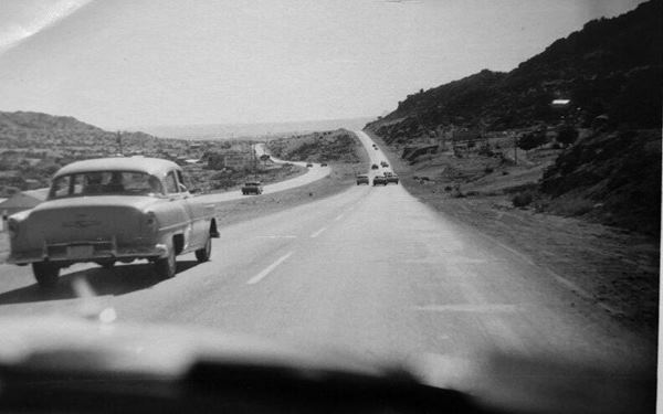 black and white photo from interior of a car: four lane highway with left side lanes skirting a hill and right side ones climbing it, cars