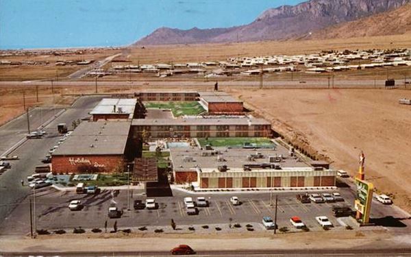 1960s aerial view of a motel, several flat roofed, two-story blocks on the property, Holiday Inn neon sign on Route 66 -lower right. Cars in parking lot
