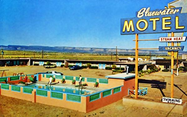 color late 1960s postcard showing a motel with cars in the background, hills and desert beyond. Deep blue sky and a swimming pool with deck chairs and people. Blue and yellow neon sign reads BLUEWATER MOTEL
