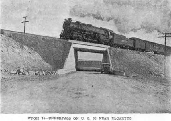 1930s black and white view of a concrete underpass, locomotive belching smoke passes overhead. Below a car. Mesa in the distance