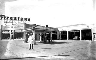 1930 Firestone Service Store black and white photo, L shaped tire store in the background, hip roof canopy as an island with pumps under it, man in the foreground