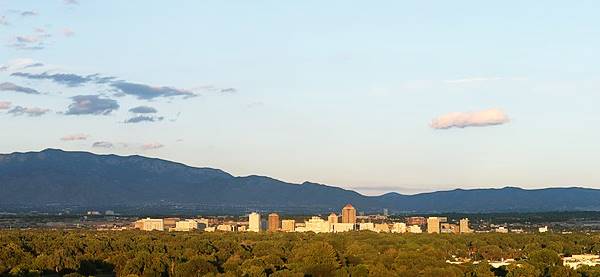 Sandia Mountains in the distance, blue sky above with some clouds, skyscrapers in the center, urban area with trees surrounds them: Albuquerque, looking east from the Rio Grande