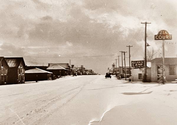 snow covered street, buildings to the left and Zia Court and Allens Camp signs on the right, car on highway