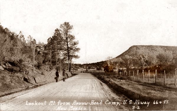Arrowhead Camp and US66 in the 1920s sepia 1920s image, dirt highway, hills on both sides, some trees, two men by the highway