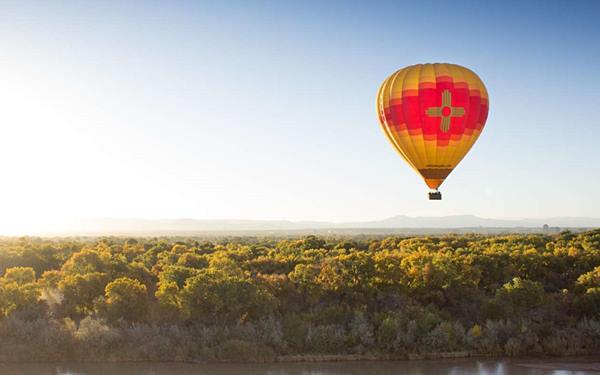 red and yellow hot air balloon floating at dusk over trees, the Rio Grande river is beneath it, mountains in the distance