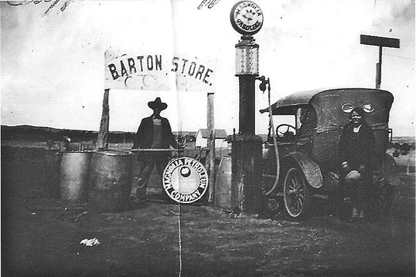 black and white picture 1920. Man by gas pump, steel drums, and car, daughter by car trunk.Magnolia gas sign and Sign above man reading BARTON STORE