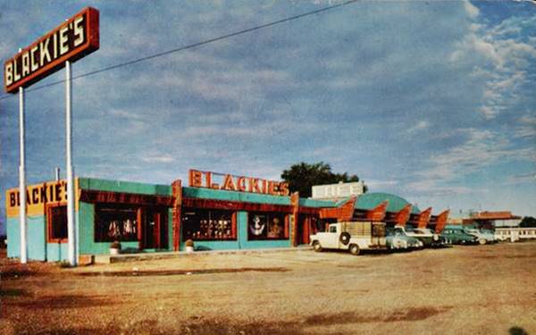 color 1950s postcard: long cafe, cars parked in front of its windows, neon sign with name, gas station to the right