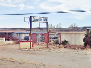 vacant single story building canopy by office with multiple-pane windows, overgrown shrubs, gravel drive. Sign reads BLUEWATER INN in blue letters on a white box shaped neon