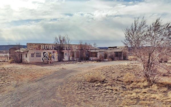 color photo. Former single story, box-shaped trading post with multiple windows and doors, grafitti and faded words reading: BOWLIN... illegible. Gravel drive, seen from Route 66