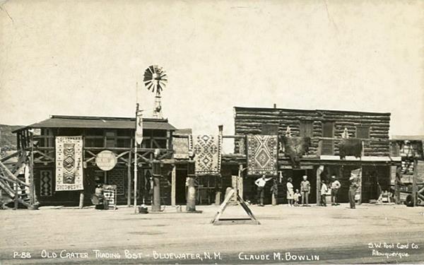 black and white picture, trading post built with logs, with rugs and bear hides hanging from the verandah and the second floor balcony. People, two Mobil gas pumps c.1930s seen from Route 66