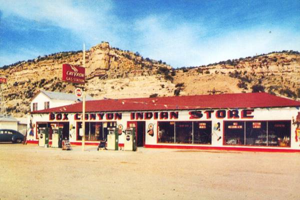 white and red single story building hip roof with 4 gas pumps in 2 islands and Chevron sign name on parapet reads BOX CANYON INDIAN STORE, cliff behind, green 1950s car to the far left
