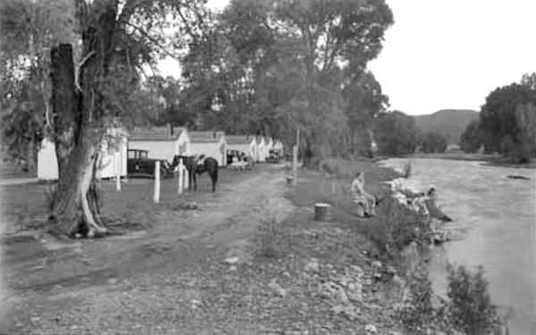 black and white 1920s, cars, horse, cabins on the left, trees; river on the right, people by it