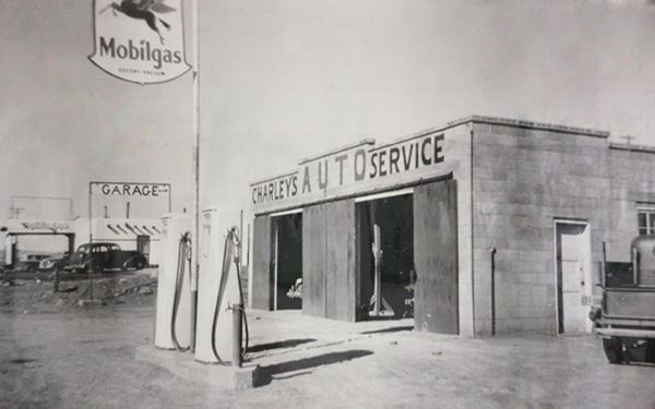 1949 black and white: garage, 2 gas pumps in front, CHARLEYS AUTO SERVICE written on parapet. Another gas station in the distance