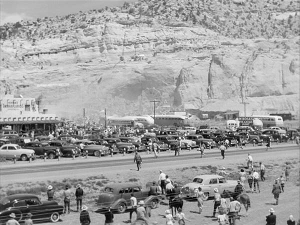 black and white still from the movie, 1951, crowds at a trading post by a cliff, cars, buses people all over the place, US 66 runs across the image