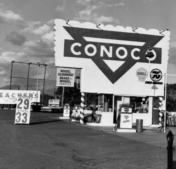 black and white view of a Conoco gas station: pumps, sign, logo, office, mountains and clouds in the distance