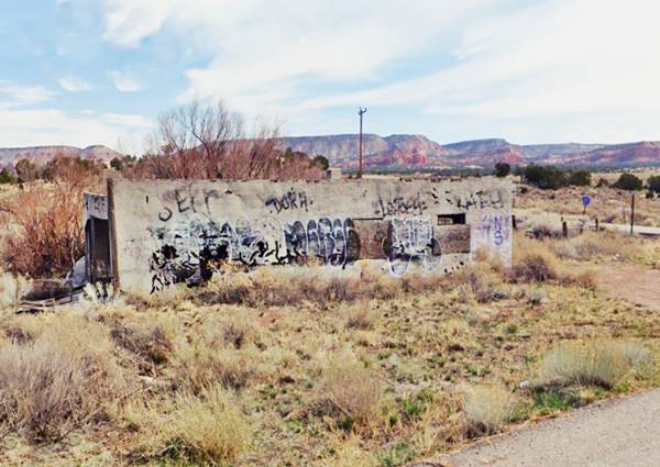 single story box shaped building, with grafitti, boarded windows by old Route 66, trees, shrubs and a mesa in the distance