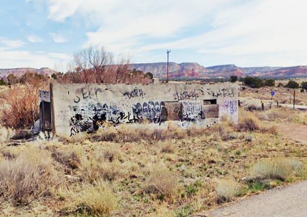 vacant building in ruins, US66 Coolidge NM in 2022 single story box shaped building, with grafitti, boarded windows by old Route 66, trees, shrubs and a mesa in the distance