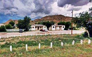 Former Dean Kirk Trading Post Manuelito NM white adobe Pueblo style buildings next to a drive beside US66, cliffs behind, trees and some cars parked