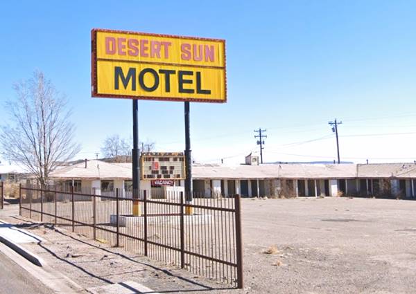 Yellow acrylic neon sign, red letters DESERT SUN and black letters: MOTEL, gable roof single floor building behind, steel fence to the left