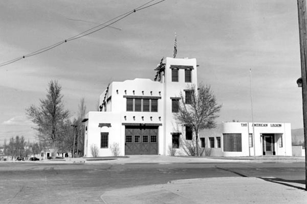 1930s black and white photo of a firehouse on a corner with Pueblo style, garage to the left, tower and American legion on the right US 66 runs in front