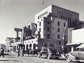Franciscan Hotel in the 1940s black and white 1940s hotel with cars, people, and highway on the left, hotel on the right. Pueblo style