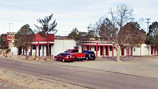 color view of a single story building, a motel, neon sign to the left, cars to the right. Seen from US 66