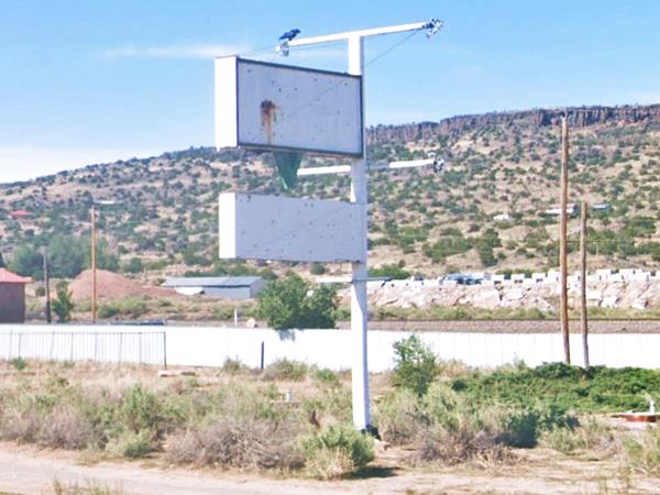 current view of a whitewashed neon sign, 2 panels and a green triangle, vacant lot with bushes, hill beyond.