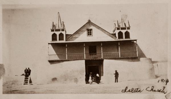 sepia 1919 whitewashed adobe church with 2 short bell towers on each side and gable roof, people.