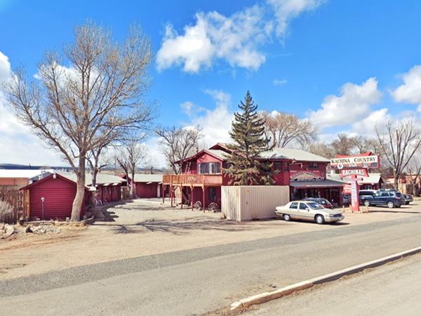 two story gable roof log cabin store by a neon sign reading KACHINA COUNTRY USA Kachinas, and partial view of log cabin units to the left, cars parked, seen from Route 66