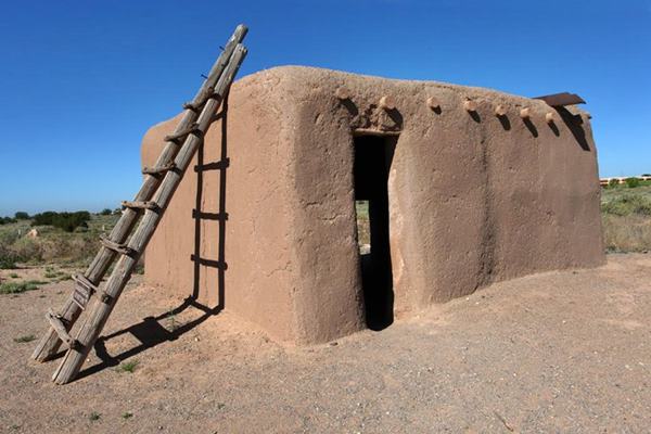 restored adobe room vigas on brown adobe wall ladder to the left, box shaped, single door, sky beyond