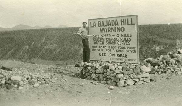 black and white 1920s Donald Crosno standing by a sign warning the dangers of the downhill road of La Bajada Hill
