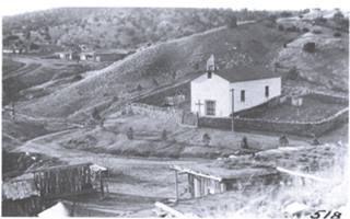 1914 view Our Lady of Light church Canyoncito black and white 1914, church, adobe, cross on its gable roof, rock wall around atrium, dirt road, some buildings on the arid hills around it