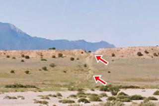 Little Cut on Route 66 desert with shrubs, a ridge with a cut in it, Sandia mountains beyond, a dirt trail runs towards the cut. Red arrows mark cut and trail
